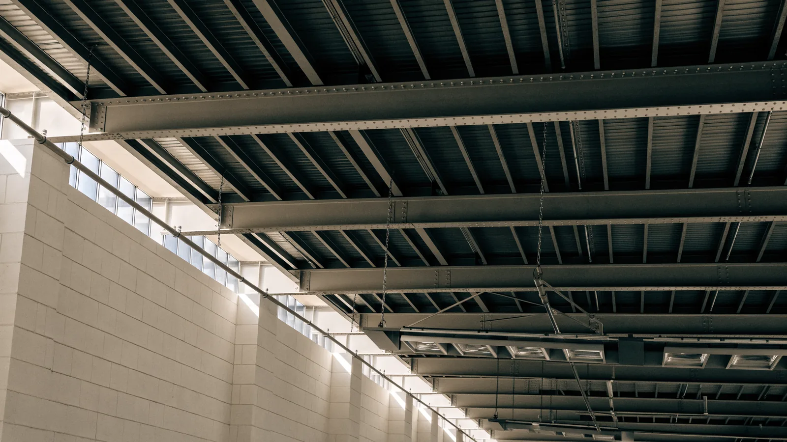 A view upward inside a warehouse to the roof structure, showing steel bar joists, metal deck, high-bay light fixtures and a sprinkler line.