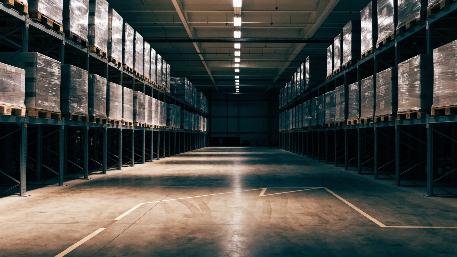 A warehouse interior with two rows of pallet racking part filled with wrapped pallets, and a clear taped staging area on the floor in front.