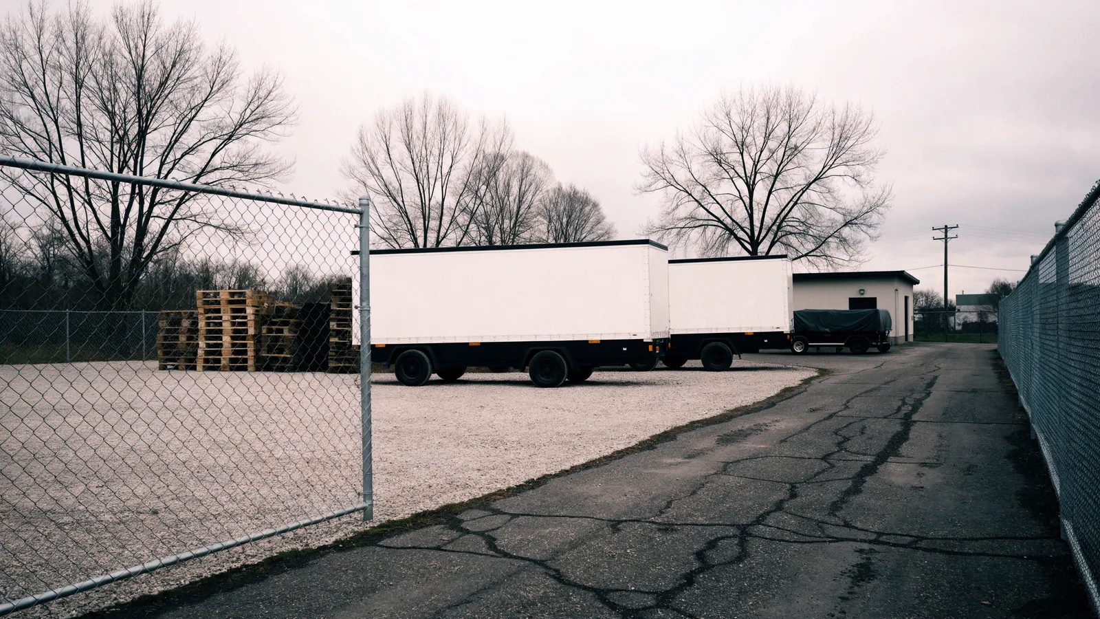 A fenced yard behind a small industrial building with two unmarked box trailers parked in line, stacked pallets and a covered equipment trailer.